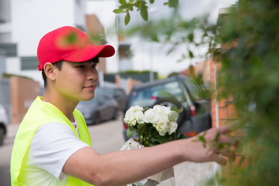 A young male flower delivery person wearing a red cap, a yellow high-visibility vest, and a white t-shirt is seen outdoors, delivering a floral gift. He is holding a bouquet of white hydrangeas wrappe