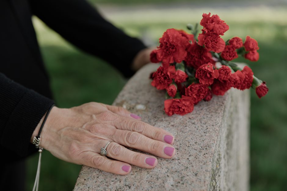 A close-up of a person's hand resting on a stone ledge, showcasing a silver ring and a black bracelet. On the ledge, there is a small bouquet of red carnations with ruffled petals, bundled together an