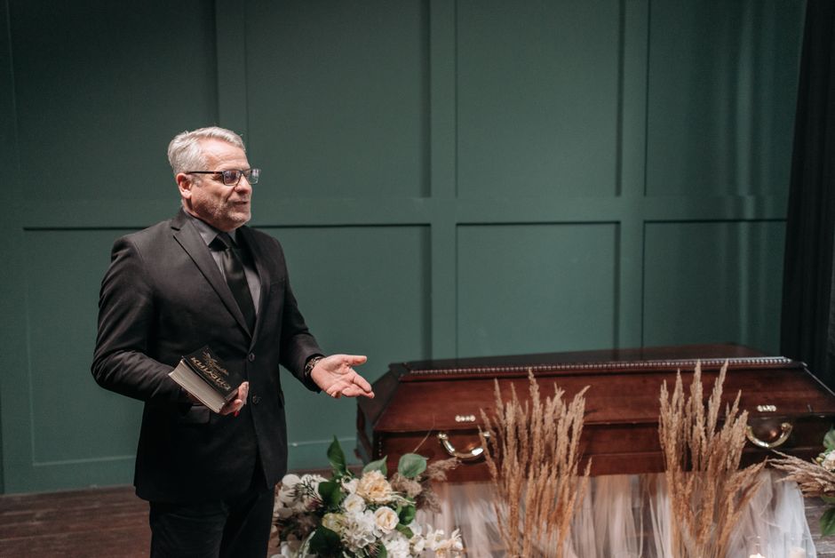 A man with grey hair and glasses, dressed in a dark suit and tie, stands in a solemn setting holding a small book or pamphlet in his left hand and gesturing with his right hand. In front of him, there
