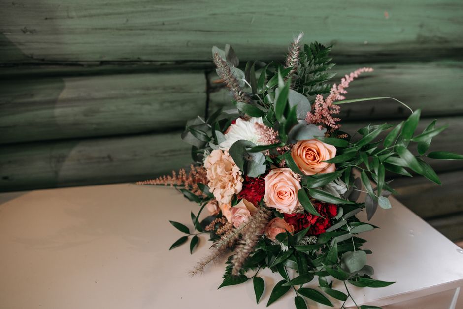 A floral bouquet featuring peach-colored roses, deep red carnations, cream-colored carnations, and pink astilbe, arranged with various types of greenery including long, slender leaves and eucalyptus.