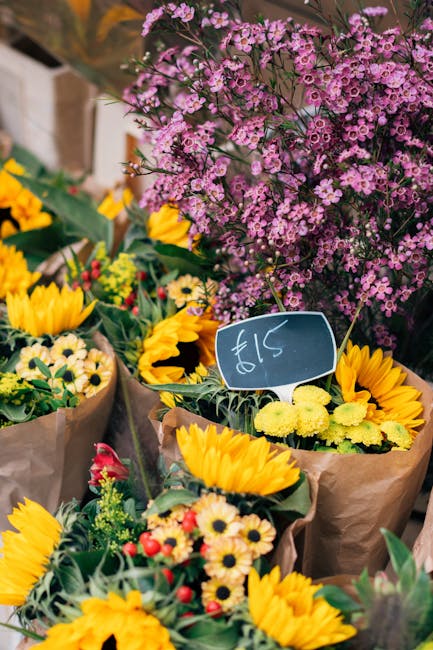 A vibrant display of fresh floral bouquets arranged with a variety of flowers including bright yellow sunflowers, small daisy-like yellow and white blooms, and red accents, wrapped in brown kraft pape