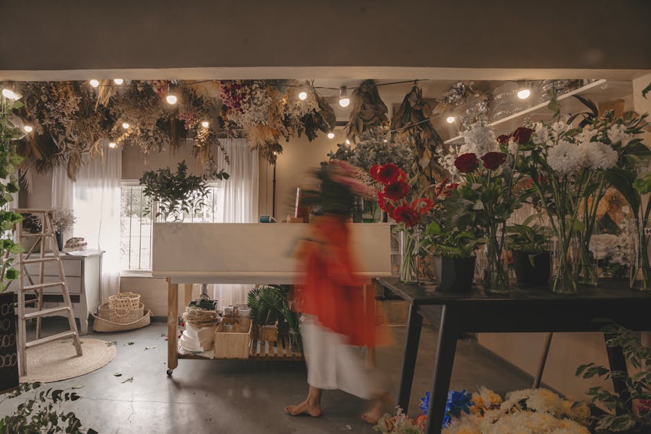 Inside a cozy flower shop with soft, warm lighting, a variety of fresh flowers in glass and black vases are arranged on a black table and nearby surfaces. The bouquet includes deep red roses, white ch