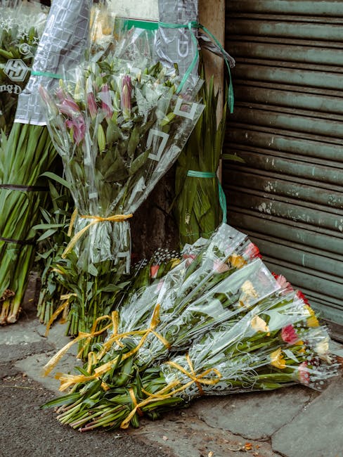 Several bouquets of fresh flowers, including pink lilies, yellow tulips, and mixed blooms, are wrapped in clear plastic and tied with yellow ribbons, leaning against a weathered wall and a closed meta