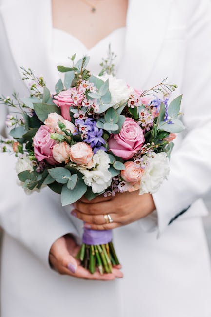 A person dressed in a white outfit is holding a floral bouquet featuring pink roses, pale peach spray roses, white lisianthus, and sprigs of purple and pink blooms, accented with green eucalyptus leav