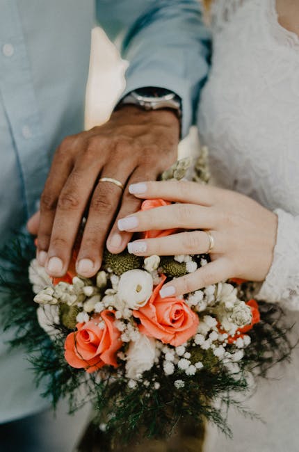 Close-up of a couple's hands resting on a vibrant floral bouquet, featuring coral roses, white ranunculus, baby's breath, and green foliage, wrapped in a soft, neutral-colored paper with a satin ribbo